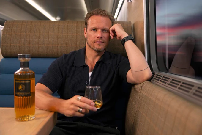 Sam Heughan in the Club Car holding a glass of whisky with a bottle of The Sassenach on the table beside him.
