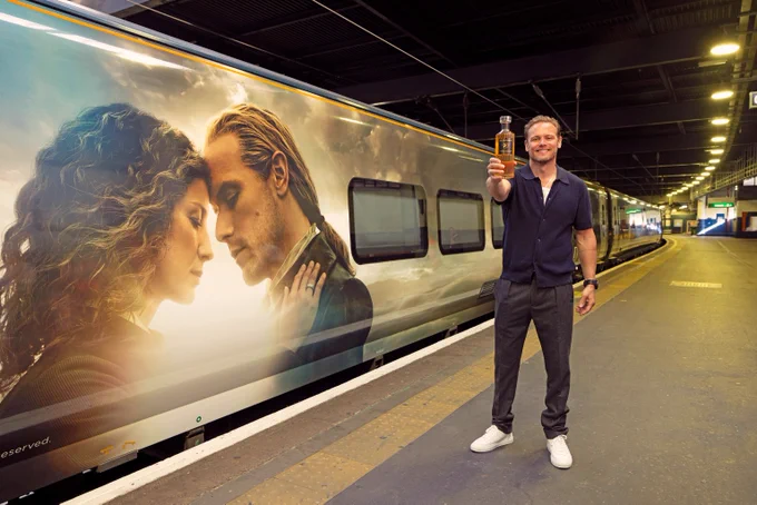 Sam Heughan standing next to the Outlander wrapped coach holding a bottle of The Sassenach whisky