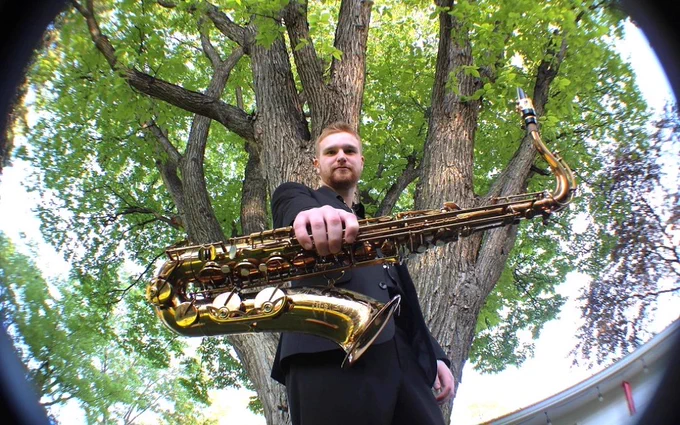 A person in a dark suit holds a saxophone towards the camera, standing in front of a large tree with abundant green leaves. The sky is clear and light can be seen filtering through the branches.