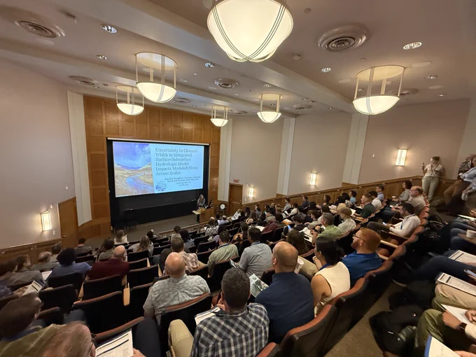 Amelia (Mia) Peeples presents her talk in a packed lecture hall. Her title slide is displayed behind her, featuring her presentation title, co-authors, a river/stream photo on the left, and the conference logo at the bottom center.