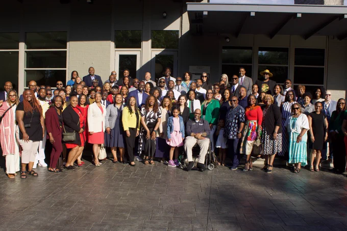 A large, diverse group of people pose together for a group photo on a paved patio outside the Governor's residence. The crowd consists of dozens of men and women dressed in business casual and colorful attire, standing in multiple rows. A man in a wheelchair sits prominently near the center front of the group, and everyone is smiling toward the camera under bright daylight.