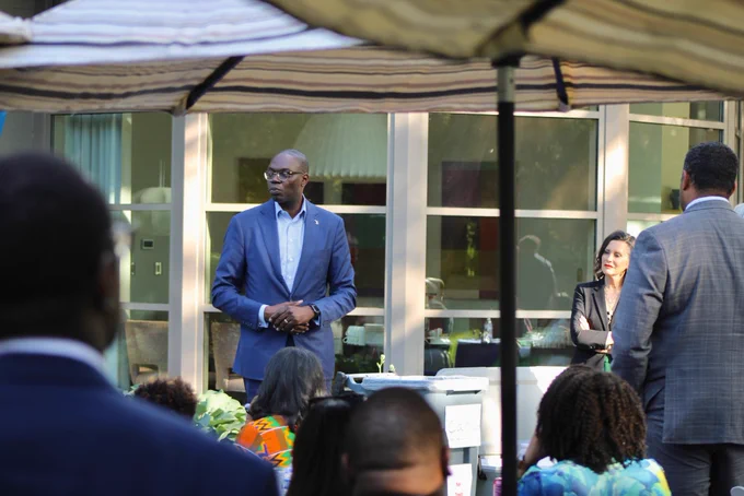 The Lieutenant Governor, wearing glasses and a blue suit jacket over a light blue button-down shirt, stands outside the Governor's residence addressing a crowd. He is looking off to the side with his hands clasped in front of him. Guests are seated in the foreground, partially obscured by a patio umbrella, and a woman stands listening in the background near large glass windows.