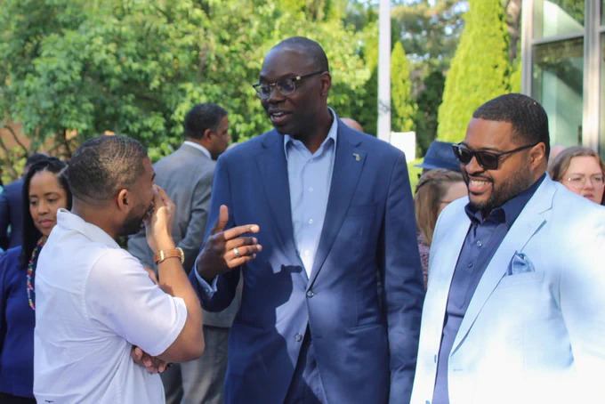 The Lieutenant Governor stands in a courtyard at the Governor's residence, engaging in a lively conversation with two men. He is wearing a blue suit jacket with a small state-shaped pin on his lapel. To his right, a man in a light blue suit and sunglasses smiles, while a man in a white shirt listens intently on his left. Lush green trees and other guests are visible in the background.