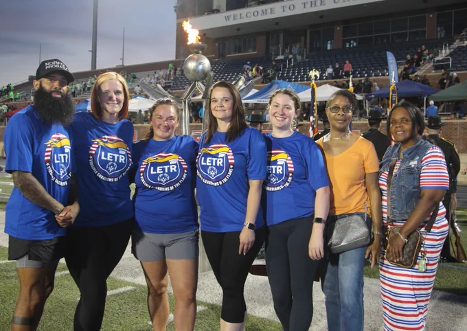 Group of seven people standing on a sports field wearing LETR event shirts with a stadium crowd in the background.