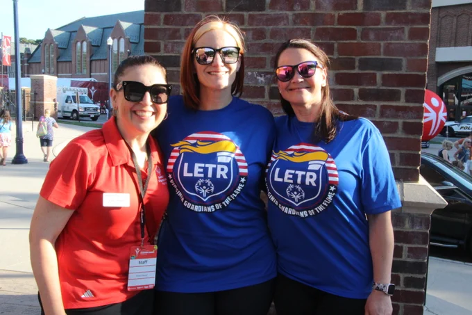 Three women standing together outdoors, two wearing blue LETR Guardians of the Flame t-shirts and one in a red shirt with a name badge.