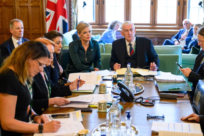 A group of people sit around a large wooden table in a meeting room with wood panelled walls and tall windows. Papers, folders, notebooks, coffee cups, and water bottles are on the table. The Speaker of the House of Commons and the President of the German Bundestag sit near the centre, looking toward the discussion. A Union Flag stands behind them.