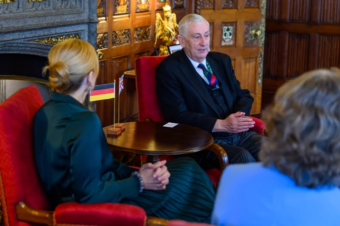 The Speaker of the House of Commons, Sir Lindsay Hoyle, and the President of the German Bundestag, Julia Klöckner, sit across from each other in red upholstered chairs in an ornate, wood panelled room, engaged in conversation. A small round table between them holds a small German flag. The Speaker speaks with hands slightly raised, while Julia Klöckner, sits turned toward them. Carved wooden walls, decorative panels, and a fireplace are in the background, with another person partially visible in the foreground.