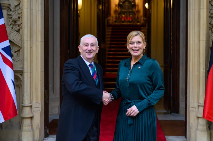 The Speaker of the House of Commons, Sir Lindsay Hoyle, and the President of the German Bundestag, Julia Klöckner, shaking hands on a red carpet outside Speaker’s House in the Palace of Westminster. They are standing in the doorway of Speaker’s House, an ornate stone building. Behind them, a staircase with a red runner leads upwards. Flags are partially visible on both sides of the entrance, including a Union Flag on the left.