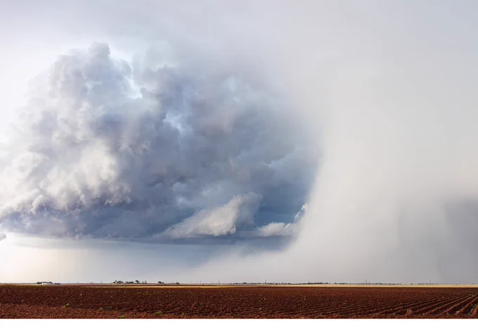 Massive dark storm cloud forming over a flat, plowed field under a gray sky.