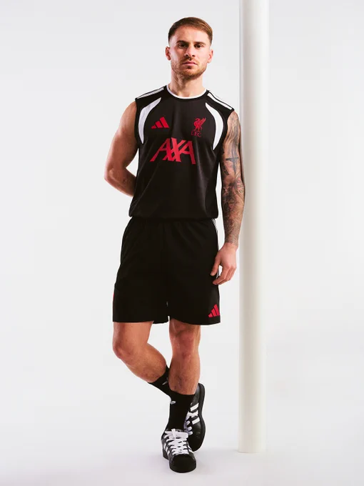 Alexis Mac Allister standing against a white backdrop wearing a black Liverpool FC sleeveless training top with red AXA sponsor and black shorts, paired with black socks and black adidas trainers, one leg crossed casually.