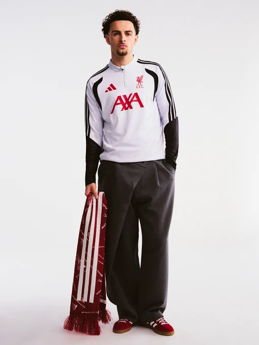Curtis Jones standing against a white backdrop wearing a white Liverpool FC training top with red AXA sponsor and black sleeve detailing, paired with loose dark trousers and red adidas trainers, holding a red striped scarf.