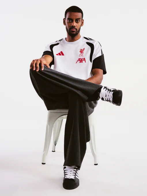 Alexander Isak seated on a simple chair wearing a white Liverpool FC training top with red AXA sponsor and black track pants, paired with black and white adidas trainers, legs crossed at the knee.