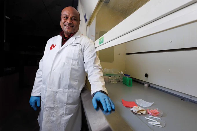 A researcher wearing a white lab coat and blue gloves stands in a laboratory next to a workbench with scientific testing materials.