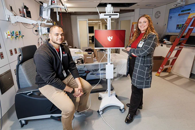 Two faculty members stand beside a mobile monitor workstation in a patient care room equipped with medical technology and equipment.
