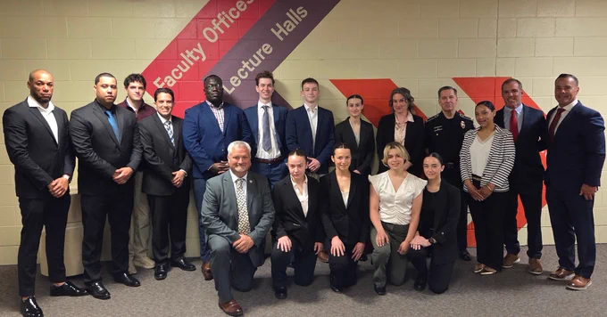 Group of professionally dressed individuals posing in front of a wall with directional signs for faculty offices and lecture halls.