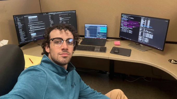 A person wearing glasses takes a selfie in an office cubicle, sitting at a desk with multiple computer monitors. The screens display various programming and software development interfaces. The workspace has a keyboard, mouse, notepad, and a pen.
