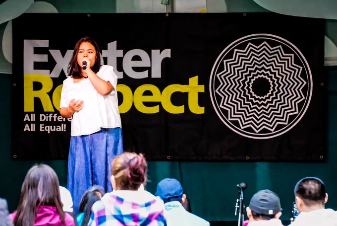 A young performer sings into a microphone on an outdoor stage. Behind them is a large “Exeter Respect” banner with the slogan “All Different All Equal,” while audience members watch from the foreground.