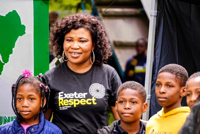 An adult and a child stand together outdoors. The adult wears a black “Exeter Respect” T-shirt with the slogan “All Different All Equal,” while the child wears a blue jacket and looks toward the camera.