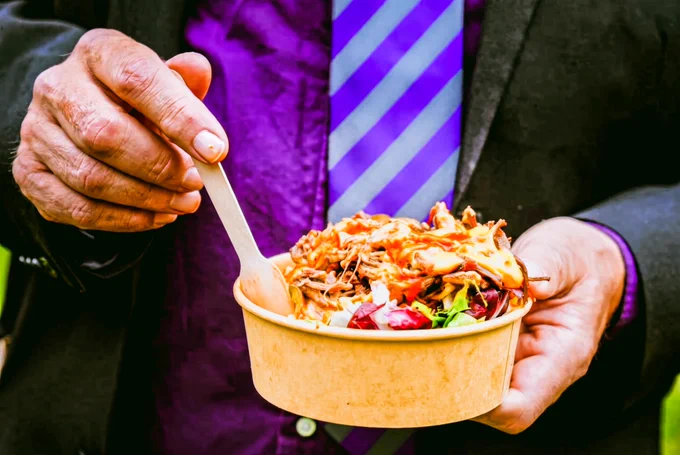 Close-up of a compostable food bowl filled with salad and shredded meat topped with sauce. Two people hold the bowl, and a person in a purple striped shirt stands behind it.