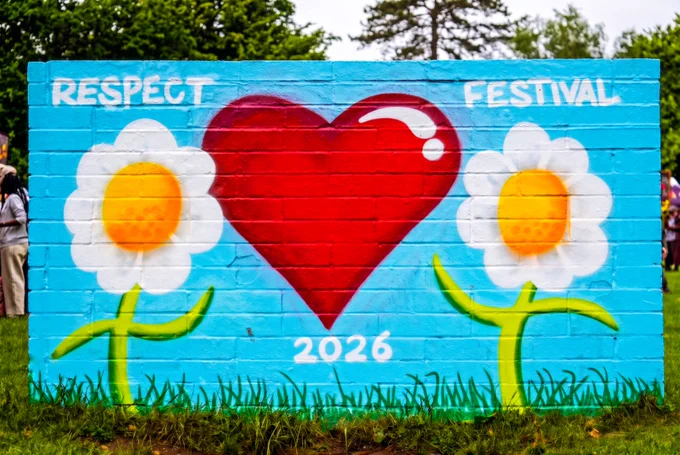 Colorful mural painted on a brick wall with the word “RESPECT” in white lettering above a large white flower with a yellow center and a red heart. Grass grows along the bottom edge, and trees are visible in the background.