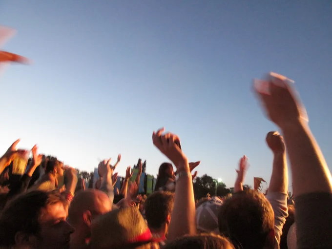 Blurred crowd at an outdoor event with many people raising their hands toward the sky in warm evening light