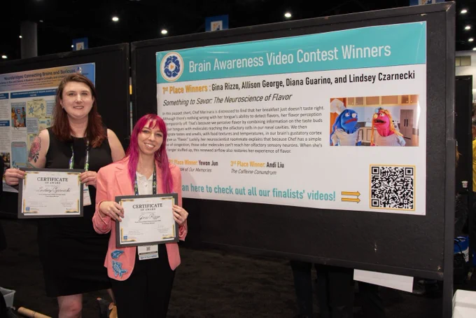 Two women proudly display their certificates in front of a poster announcing the winners of the Brain Awareness Video Contest. The mood is celebratory and professional.