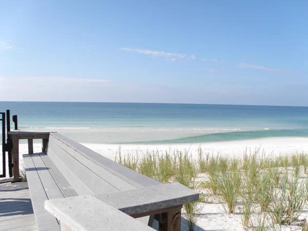 Wooden boardwalk bench overlooking a calm ocean with white sandy beach and sparse dune grass under a clear blue sky.