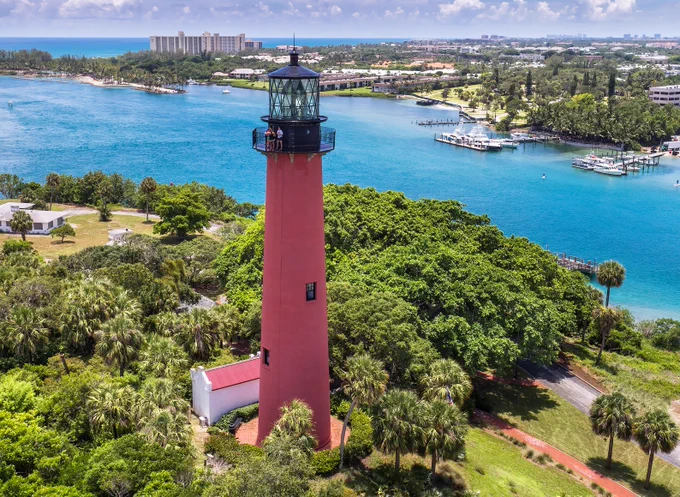 Tall red lighthouse surrounded by lush green trees near a vibrant blue coastal waterway under a partly cloudy sky.