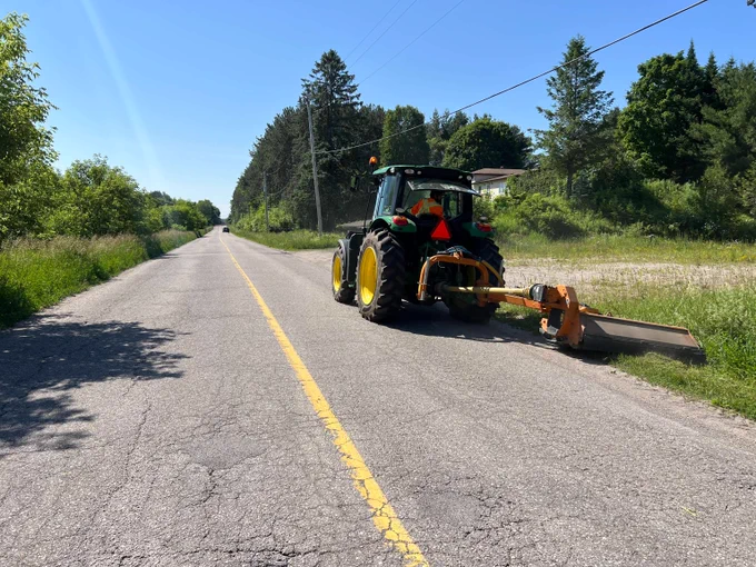 Green tractor mowing grass along a rural roadside in the Township of Tiny on a sunny day with clear blue sky.