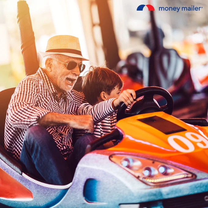 Elderly man wearing sunglasses and a hat joyfully rides a bumper car with a young boy steering the wheel.