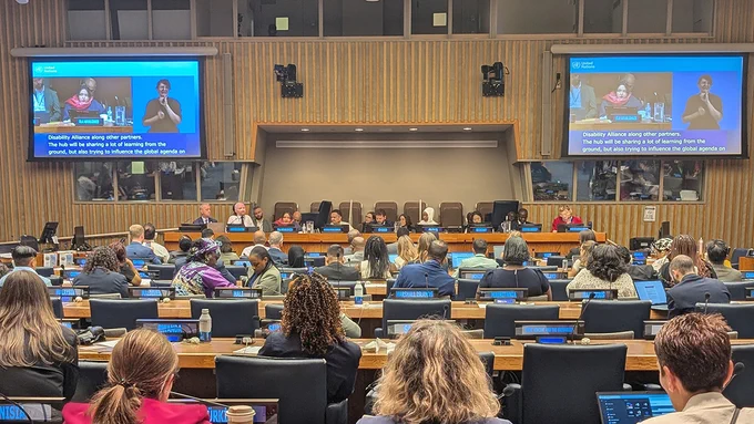 A large UN conference hall filled with delegates seated at desks with microphones and laptops. At the front, a panel speaks beneath large screens showing live captions and a sign language interpreter. Nameplates identify countries, and rows of attendees listen while some take notes or follow the session on screens.