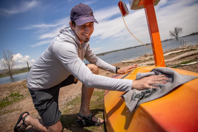 Man smiling while drying an orange kayak with a grey towel with a lake in the background.