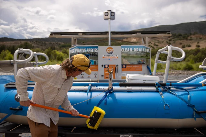 Man cleaning a blue water raft with a yellow brush near a cleaning station to help protect against aquatic invasive species.