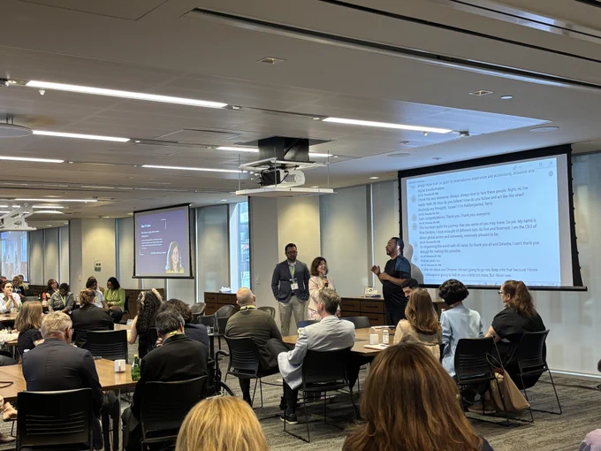Giuseppina (Pina) D'Intino speaks to a room of accessibility professionals who are all seated at tables. To the right of her is an ASL interpreter.
