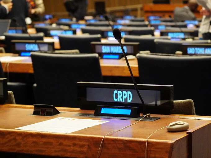 A conference room at the United Nations with rows of desks and microphones. In the foreground, a desk has a sign reading “CRPD,” along with a headset, cables, and a document. In the background, multiple seats and nameplates are visible, softly lit, with a few people standing and moving around.