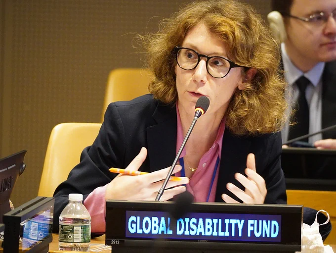 Natalia Mattioli with blond hair and glasses speaks into a microphone at a United Nations meeting table. She wears a dark blazer and holds a pen while gesturing. In front, a sign reads “GLOBAL DISABILITY FUND.” A water bottle and papers sit nearby, with another attendee blurred in the background.