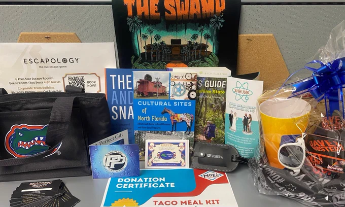 A collection of available prizes, including a U F lunchbox, U F flag, U F luggage tag, Escapology gift certificate, Moe’s gift certificate, three books from the U F Bookstore, a Dave and Buster’s gift baskets and gift certificate, a Shrunk 3 D gift card and pamphlet, Bragging Rights Amusements gift cards, a Park Place Car Wash gift card, and Adam’s Rib Co. gift cards.