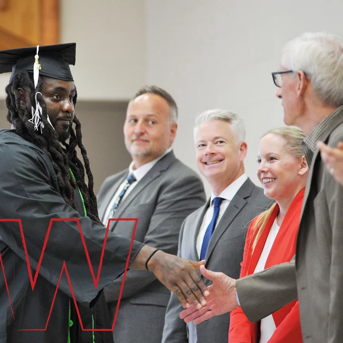 A recent graduate of the Prison Education Initiative wearing a cap and gown and shaking hands with Governor Tony Evers.