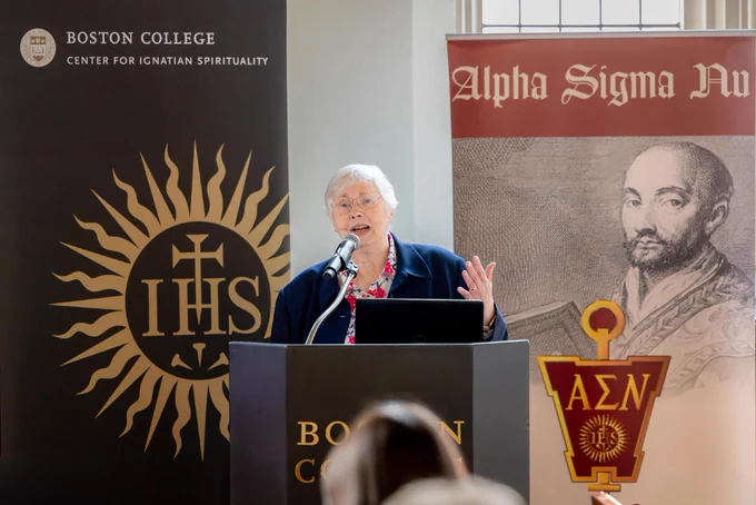 Sr. Bartunek speaks at a podium with the words "Boston College" on it. Behind them are banners for Boston College Center for Ignatian Spirituality and Alpha Sigma Nu, featuring a historic image.