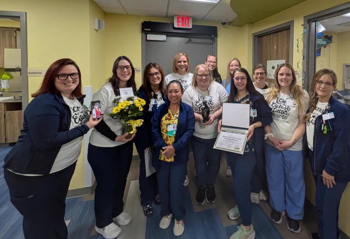 A group of healthcare professionals, some holding a bouquet of yellow flowers and an award certificate, are smiling in a hospital corridor. 