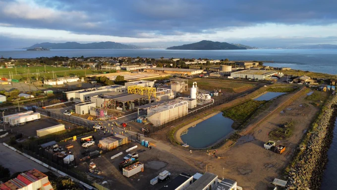 Aerial view of the Treasure Island Water Resource Recovery Facility in San Francisco, California, showing treatment infrastructure, operational buildings and waterfront surroundings. The sustainable water infrastructure project sits along San Francisco Bay with surrounding community development visible in the background.