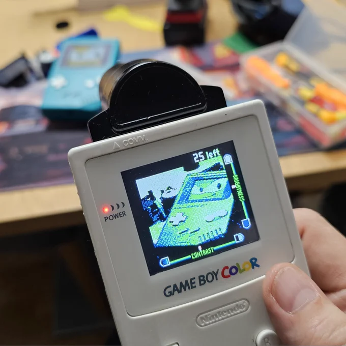 Ein Game Boy mit Game Boy Kamera. Am Display ist ein weiterer Game Boy zu sehen. Offensichtlich wird mit der Game Boy Kamera ein am Tisch liegender Game Boy fotografiert. 