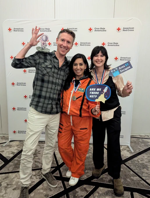 Three individuals are standing in front of a backdrop with "American Red Cross" and "Cruz Roja Americana" logos. One person is holding a small trophy, another is dressed in an orange astronaut costume, and the third holds signs saying "Are We There Yet?" and "Area 51 Escape." They are smiling and appear to be at an event.