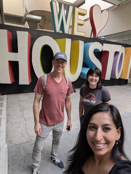 Three people smile in front of a large sign that reads "We Love Houston.