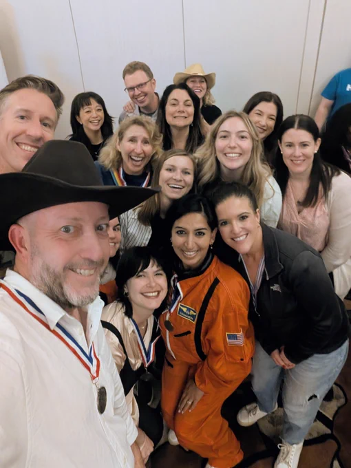 A group of smiling people gathers closely for a selfie. One individual in the front is wearing a cowboy hat, and another is dressed in an orange NASA astronaut suit. Some attendees are wearing medals around their necks. The background shows a plain wall.