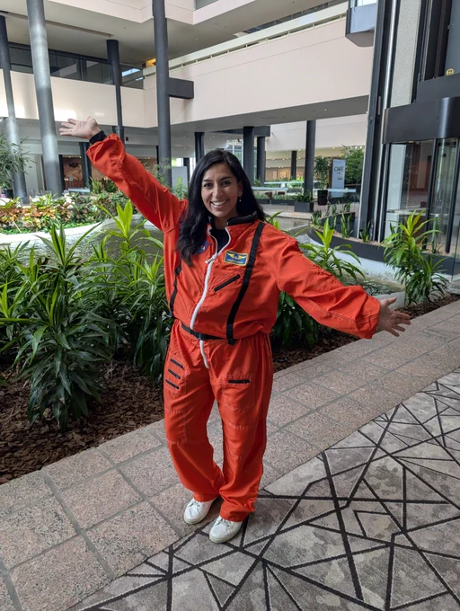 An individual standing with arms outstretched, wearing a bright orange NASA jumpsuit, is in an indoor space with plants and modern architecture.