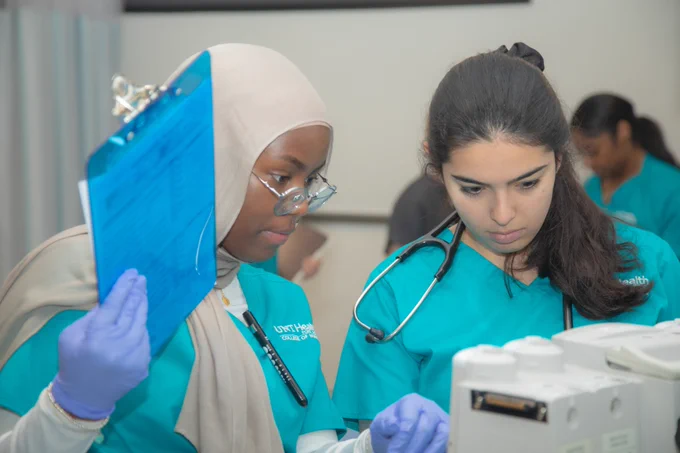 Two students in teal uniforms and gloves focus on a task. One is wearing a stethoscope and the other holds a clipboard. They are in a clinical setting with medical equipment nearby.