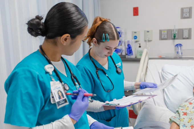 Two students in teal scrubs and gloves are examining documents at a patient's bedside in a hospital room.