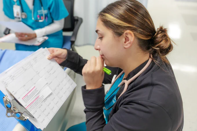 A student wearing a stethoscope is seated at a desk, closely examining a document with medical charts. Another person, in medical attire, sits nearby holding papers.