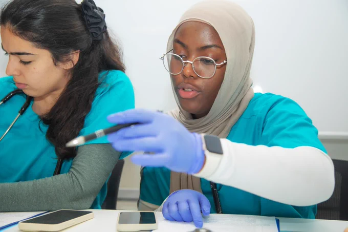 Two students are seated at a table. One person is wearing a headscarf and gloves, holding a pen and pointing at a document. Both wear turquoise scrubs and have stethoscopes. Cell phones and documents are on the table.